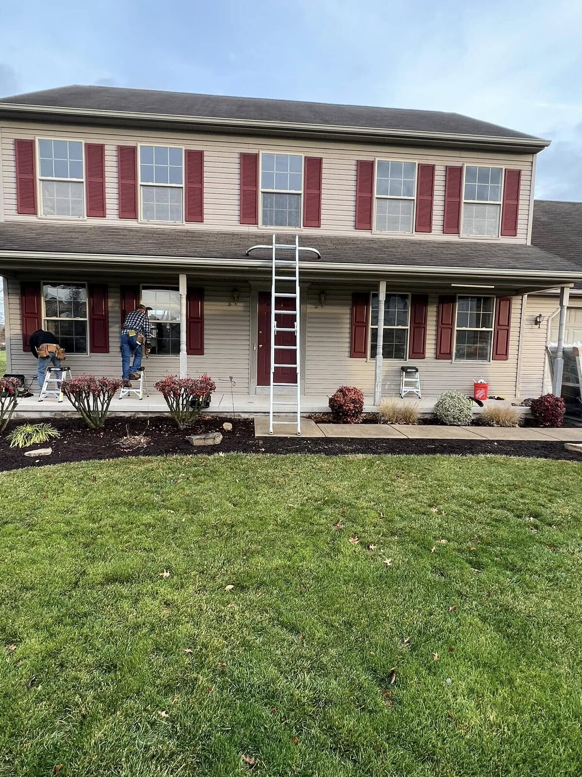 Two installers replacing front windows on a large tan colonial home with red shutters in York, Pennsylvania